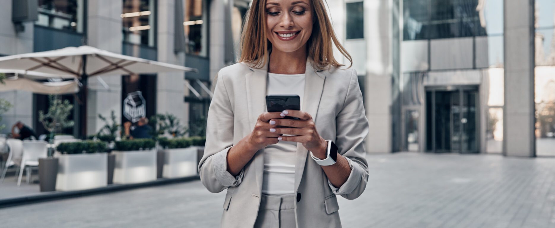 Woman in suit using smart phone and smiling while standing outdoors.